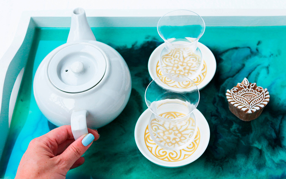 Artesanato de bandeja feita em resina epóxi, na cor azul, com duas taças em cima e um bule sendo segurado por uma mão feminina.